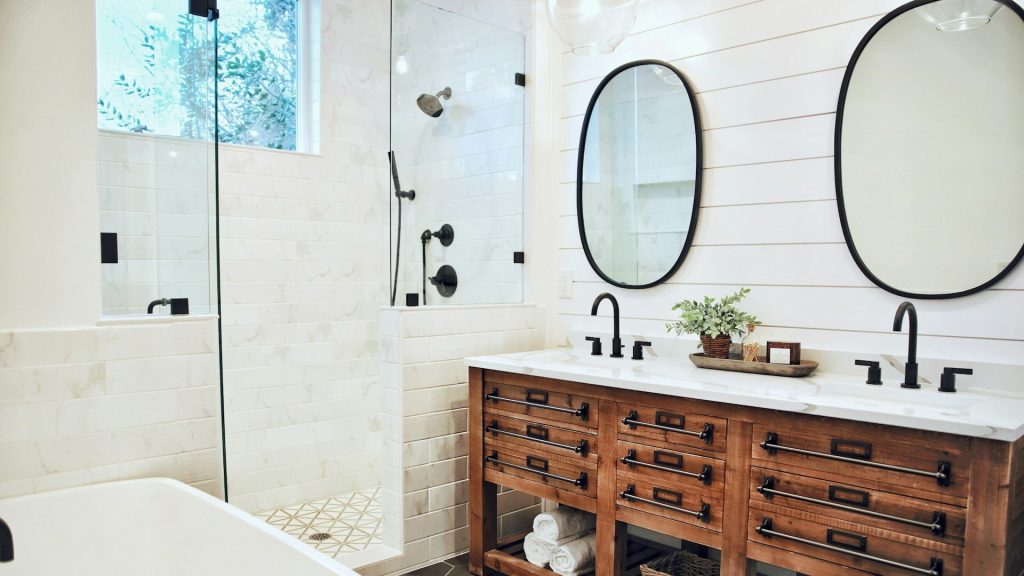 Landelijke badkamer met houten ladekast, zwarte kranen, witte wandtegels en dubbele spiegels.
